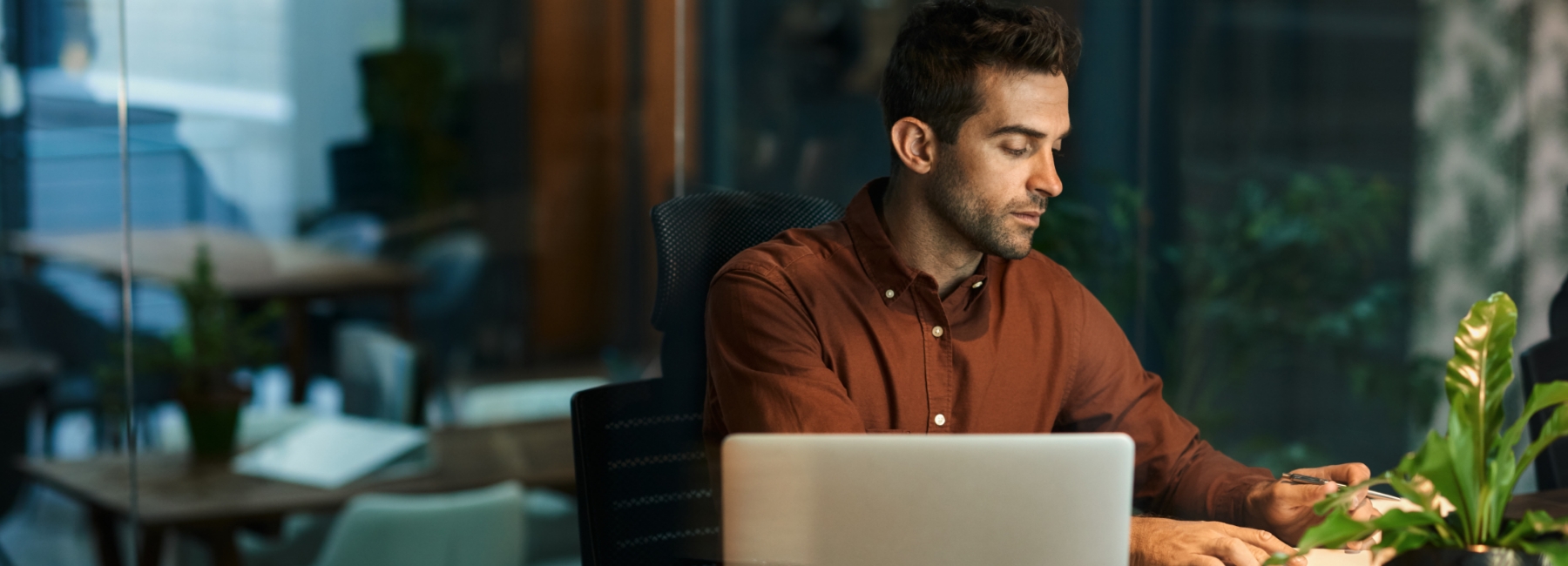 A man working in a modern office setting; he is seated at a wooden desk with an open laptop in front of him. He is wearing a brown shirt and appears to be focused on his work, using a pen to jot down notes in a notebook. The desk also has a pair of glasses and a small potted plant. The background features glass walls, offering a glimpse into a contemporary office space with additional tables, chairs, and greenery, suggesting a productive and well-designed work environment. The overall ambiance is professional and calm.