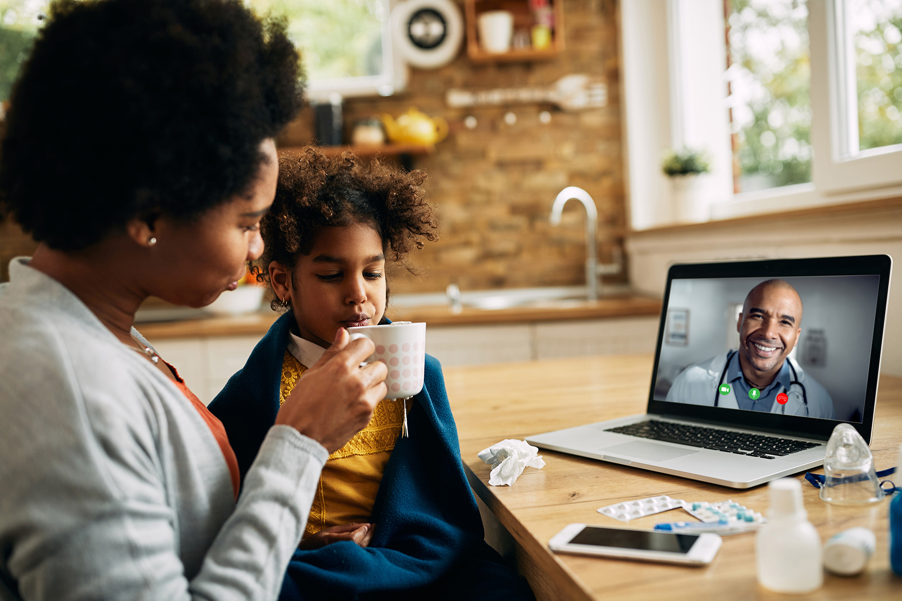 A mother is giving a cup of tea to her sick young daughter while having a video call with a doctor from home. The video call screen shows the doctor, indicating a remote medical consultation.