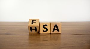 A wooden cube on a table is being turned to change the visible text from "FSA" (Flexible Spending Account) to "HSA" (Health Savings Account). The background is a simple white, emphasizing the transition between the two healthcare financial concepts.