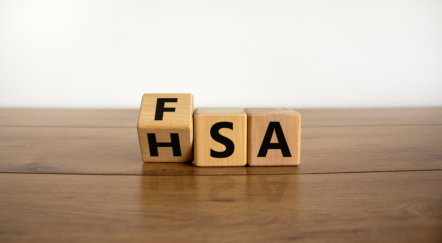 A wooden cube on a table is being turned to change the visible text from "FSA" (Flexible Spending Account) to "HSA" (Health Savings Account). The background is a simple white, emphasizing the transition between the two healthcare financial concepts.