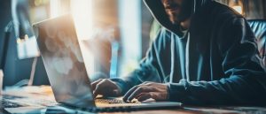 A person in a dark hoodie working on a laptop, symbolizing cyber fraud, online scams, stolen data, and digital theft. The background is dark and features binary code and digital symbols.
