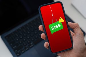 A smartphone screen displays a text message scam alert, indicating a "smishing" concept. The message contains a warning about a suspicious link or fraudulent activity. The background is neutral, highlighting the phone and the scam notification.