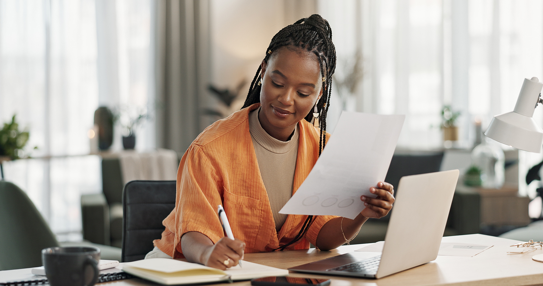 A woman sits at a desk in a brightly lit room, holding a piece of paper in one hand and writing in a notebook with the other. She wears an orange jacket and works on a laptop, surrounded by a cup of coffee, a lamp, and office supplies.