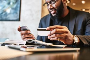 A businessman is sitting in a café, holding a mobile phone and a credit card. He appears to be checking his account, with a focused expression on his face. The café setting includes tables and a warm, inviting atmosphere.