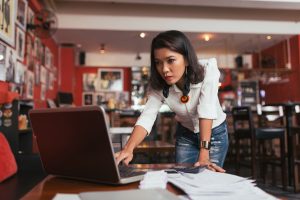 A concerned businesswoman is at a desk. She is holding a pen and looking at documents or paperwork in front of her. The background is a typical office setting with a computer and office supplies.