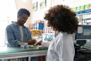 A man is making a payment while shopping for medicine at a pharmacy. The background includes blurred store shelves in a shopping environment.
