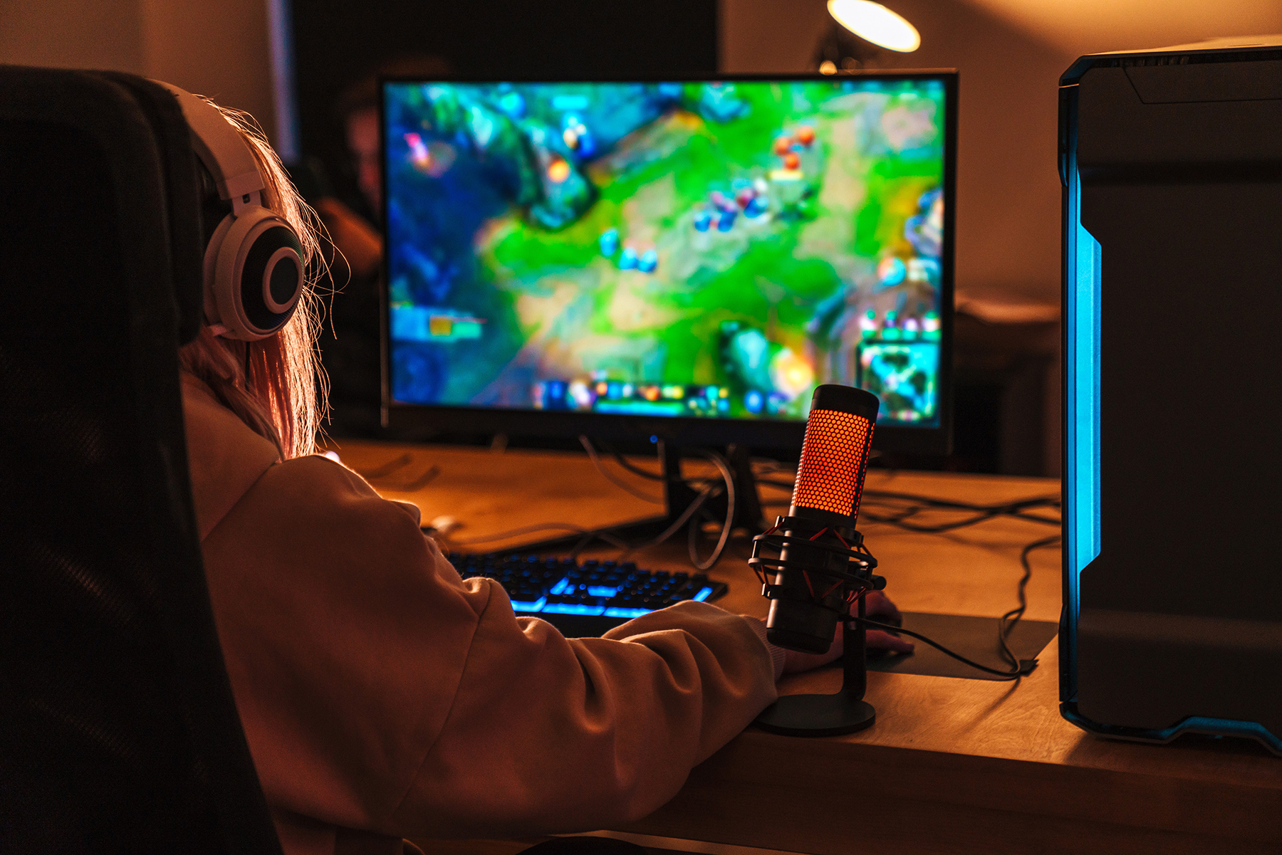 The image shows the back of a girl playing a video game on a personal computer. She is seated at a desk. The screen displays vibrant, colorful game graphics.