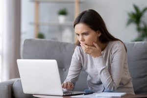 A shocked and stressed young woman is sitting at a desk, reading bad online news on a laptop screen. She appears confused and panicked, frustrated by a computer problem, mistake, or negative social media message.
