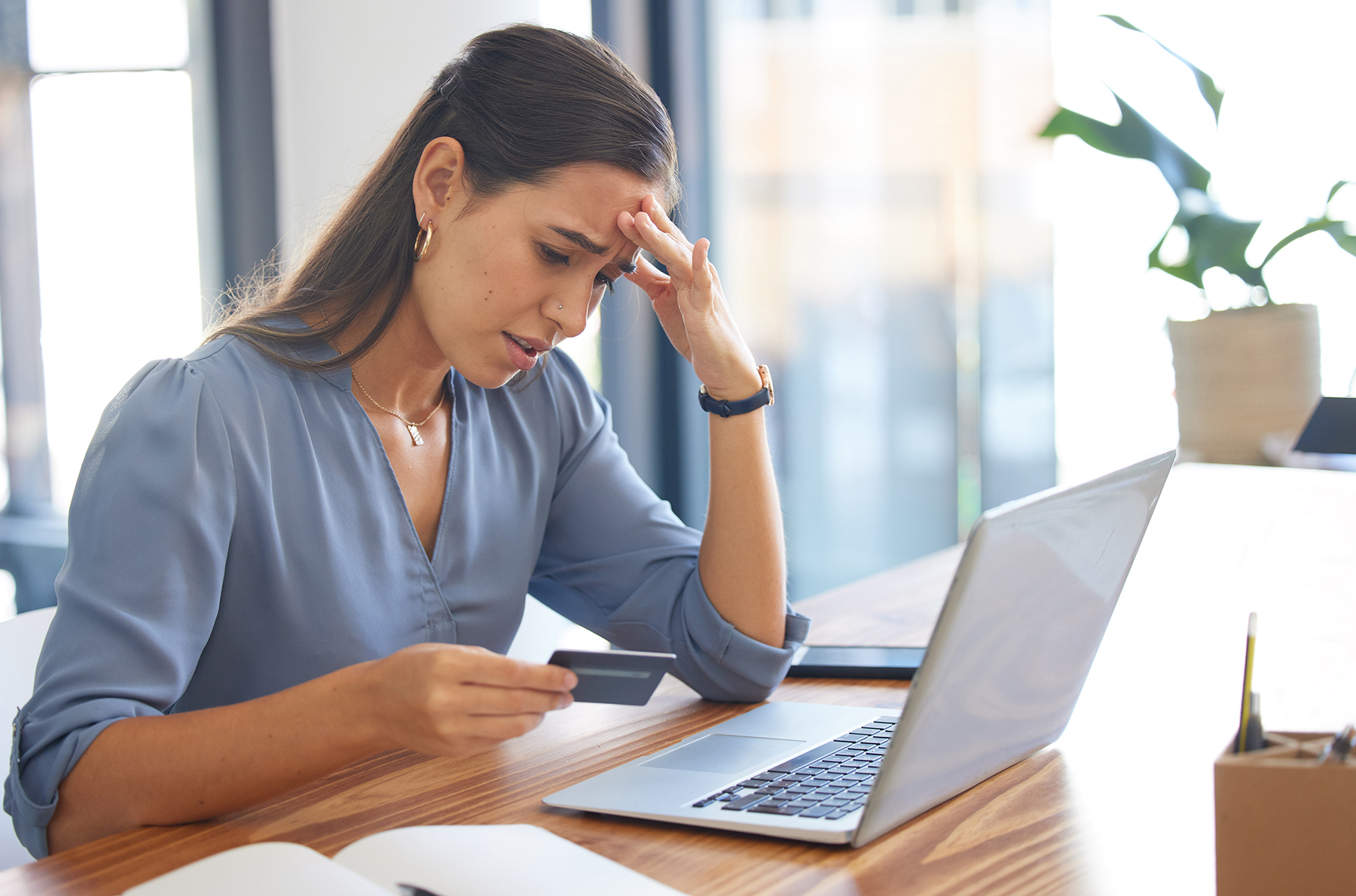 Young woman sitting at a desk, holding a credit card and looking worried or stressed as she views her laptop screen. The setting appears to be a modern office or home workspace with large windows in the background.