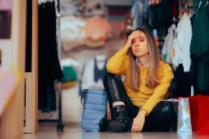A tired woman, appearing overwhelmed, is sitting on the floor of a store during a shopping spree. She looks unhappy and exhausted, resting amid shopping bags and clothing racks.