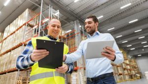 A warehouse worker and a businessman are standing together in a large warehouse. The worker is holding a clipboard, while the businessman is using a tablet. The background shows rows of shelves filled with boxes and goods, emphasizing the scale of the wholesale logistics business.
