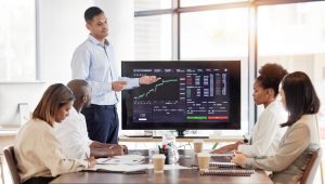 A group of business people is gathered around a large screen displaying a stock market dashboard. The screen shows various graphs, charts, and data analytics related to trading, finance, and investment. A coach or presenter is standing by the screen, explaining the information.