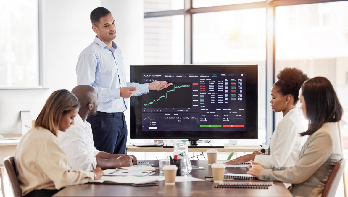 A group of business people is gathered around a large screen displaying a stock market dashboard. The screen shows various graphs, charts, and data analytics related to trading, finance, and investment. A coach or presenter is standing by the screen, explaining the information.