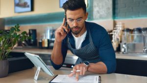 A business owner is on a phone call while reviewing paperwork in a restaurant or café setting. The scene includes a supply chain checklist or inventory documents.