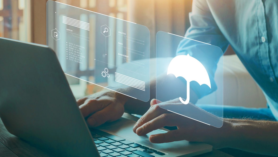 A person is typing on a laptop with transparent digital screens projected above the keyboard. One screen displays an umbrella icon, symbolizing insurance or protection, while the others show various data and management-related icons.