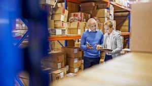 A man and a woman are inspecting inventory in a large distribution warehouse. They are looking at items on a shelf. The background shows tall shelves filled with boxes.