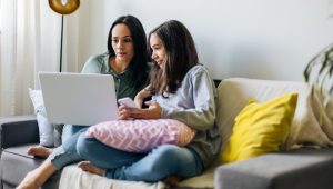 A woman and a young girl are sitting on a couch together, both focused on a laptop screen. The woman is holding the laptop while the girl, holding a smartphone, leans in closely, engaged in what they are viewing. The scene is cozy and relaxed, with soft lighting and cushions around them.