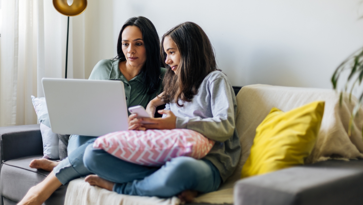 A woman and a young girl are sitting on a couch together, both focused on a laptop screen. The woman is holding the laptop while the girl, holding a smartphone, leans in closely, engaged in what they are viewing. The scene is cozy and relaxed, with soft lighting and cushions around them.