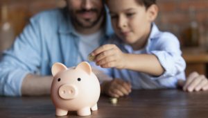 A child is placing a coin into a pink piggy bank while an adult watches closely.