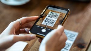 A person is using a smartphone to scan a QR code on a wooden table. The phone's screen displays the QR code in the camera app, and another QR code is visible on the table.