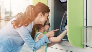 A mother and her young daughter smile as they use an ATM together, with the mother pointing at the screen while the daughter’s hand is on the machine.