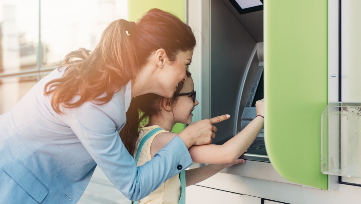 A mother and her young daughter smile as they use an ATM together, with the mother pointing at the screen while the daughter’s hand is on the machine.