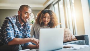A smiling couple works together on a laptop in a bright, cozy living room.
