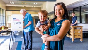 A smiling woman holding a baby stands in a voting center, both wearing "I Voted" stickers. In the background, an older man is casting his vote at a booth with a "VOTE" sign featuring an American flag.