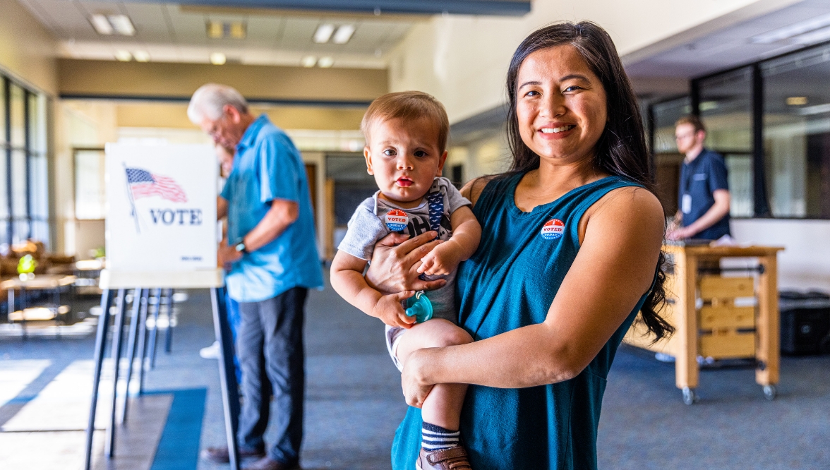 A smiling woman holding a baby stands in a voting center, both wearing "I Voted" stickers. In the background, an older man is casting his vote at a booth with a "VOTE" sign featuring an American flag.