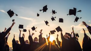 A group of graduates in black gowns celebrate by throwing their caps into the air against a backdrop of a setting sun and clear sky. The scene captures the joy and excitement of graduation day.