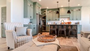 A modern kitchen and living room with sage green cabinets, a dark wood island with barstools, and stainless steel appliances. The living area includes a striped beige armchair, cushioned ottoman, and light hardwood flooring.