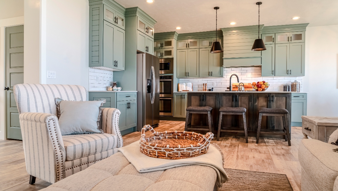A modern kitchen and living room with sage green cabinets, a dark wood island with barstools, and stainless steel appliances. The living area includes a striped beige armchair, cushioned ottoman, and light hardwood flooring.