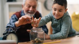 A joyful elderly man and a young boy smile as they drop coins into a glass jar. The man gently guides the boy's hand, showing him how to save money. They are seated at a wooden table, with coins scattered around and a pencil nearby.