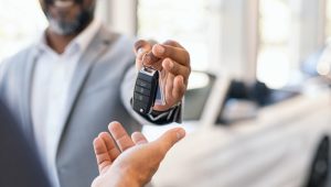 A car salesperson hands over a key to a customer in an auto showroom, with a blurred luxury car visible in the background.