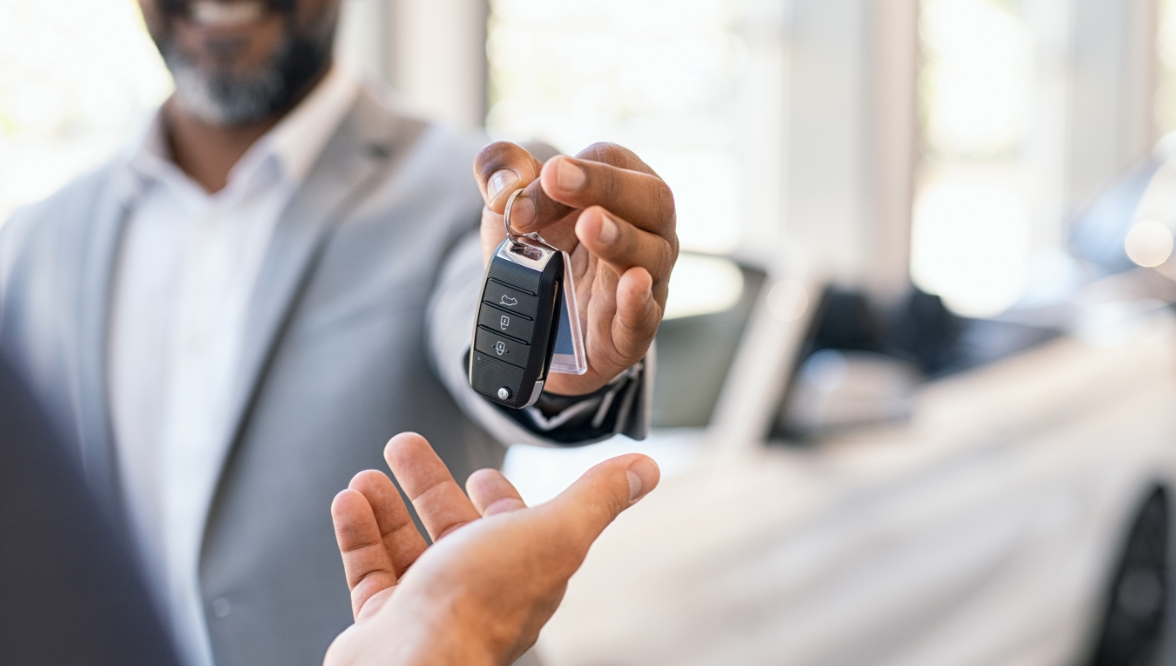 A car salesperson hands over a key to a customer in an auto showroom, with a blurred luxury car visible in the background.