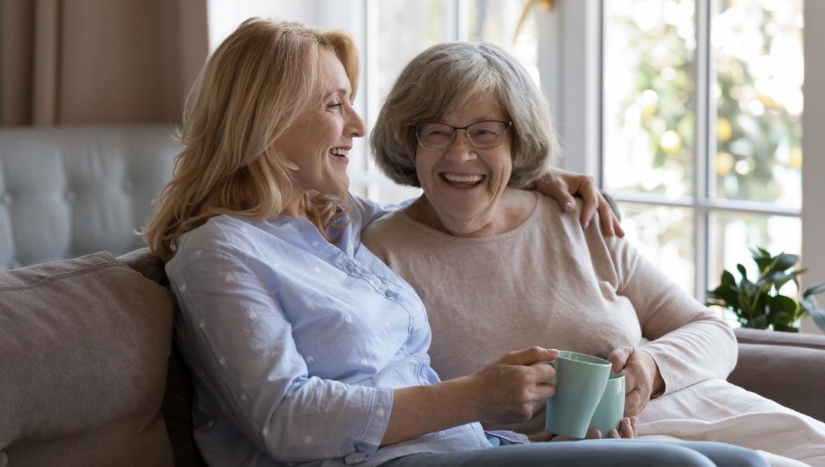 Two women, one younger and one older, sit together on a couch, smiling and laughing while holding mugs in a cozy living room setting.