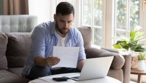 A man with a beard is sitting on a couch at home, reviewing a document while working on his laptop with a calculator on the table beside him.