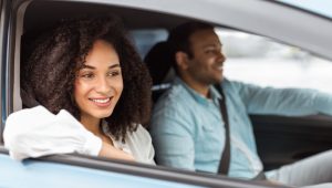 A smiling woman sits in the passenger seat of a car with the window down, while a man drives beside her, both enjoying the ride.