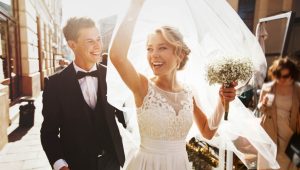 A smiling bride holding a bouquet and veil flowing behind her, walking joyfully with her groom in a sunny outdoor setting.