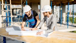 Two men wearing hard hats are reviewing blueprints at a construction site, one in blue overalls and the other in a gray suit. They are working at a table with a laptop, surrounded by scaffolding and concrete structures in progress.