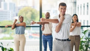 A group of four coworkers is stretching together in an office with large windows and a cityscape in the background. The man in the foreground smiles while stretching his arm across his chest, and the others behind him follow the same stretch.