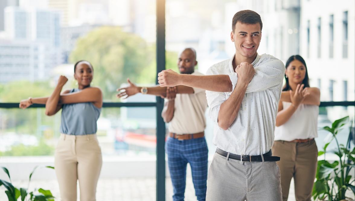 A group of four coworkers is stretching together in an office with large windows and a cityscape in the background. The man in the foreground smiles while stretching his arm across his chest, and the others behind him follow the same stretch.