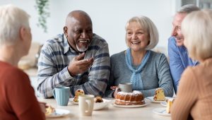 A group of older adults is sitting together at a table, enjoying coffee, cake, and each other's company. They are laughing and having a pleasant conversation in a bright, relaxed setting.