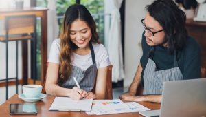 Two small business owners in striped aprons sit together at a wooden table with a laptop and coffee cup, looking at graphs while smiling. The female is writing down notes on a clipboard while the male looks at what she is writing.