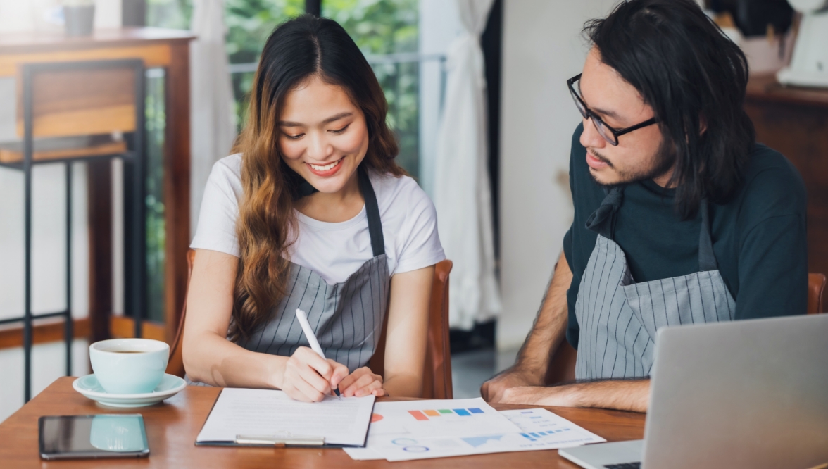 Two small business owners in striped aprons sit together at a wooden table with a laptop and coffee cup, looking at graphs while smiling. The female is writing down notes on a clipboard while the male looks at what she is writing.