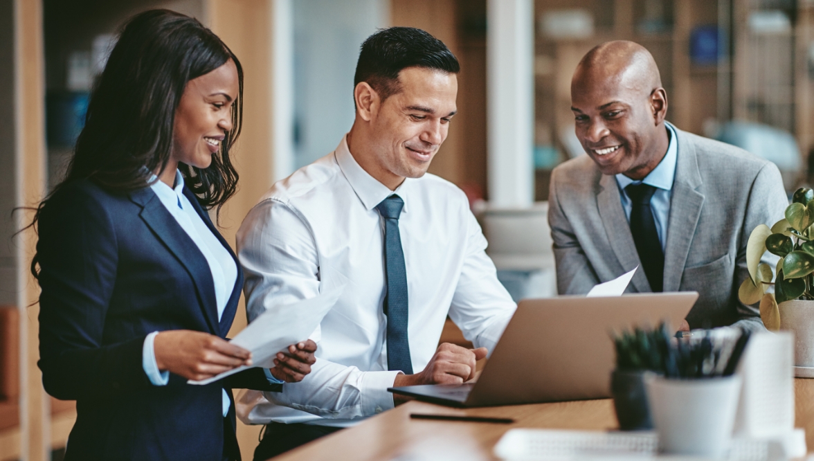 A group of three professionals, two men and one woman, are collaborating in an office setting. The woman, dressed in a navy blue blazer, holds papers while the man in the middle, wearing a white shirt and tie, works on a laptop. The man on the right, in a light gray suit, smiles while looking at the screen.