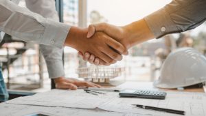 Close up of two people shaking hands over a table. On the table you can see construction plans, a hard hat, a tablet, and a calculator.