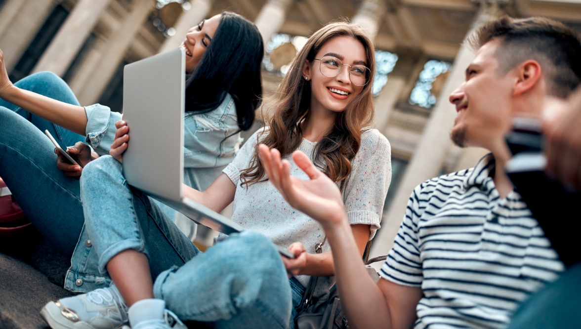 Two young people sit on the steps outside a school building, engaged in conversation. One girl smiles while holding a laptop, with other students visible in the background.