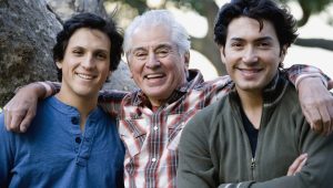 Three men from different generations smiling outdoors, with their arms around each other. The older man in the center has white hair, while the two younger men have dark hair.