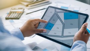 A person holds a tablet displaying a property plot map with numbered sections, focusing on plot 230. A calculator, pen, and additional documents are visible in the background.